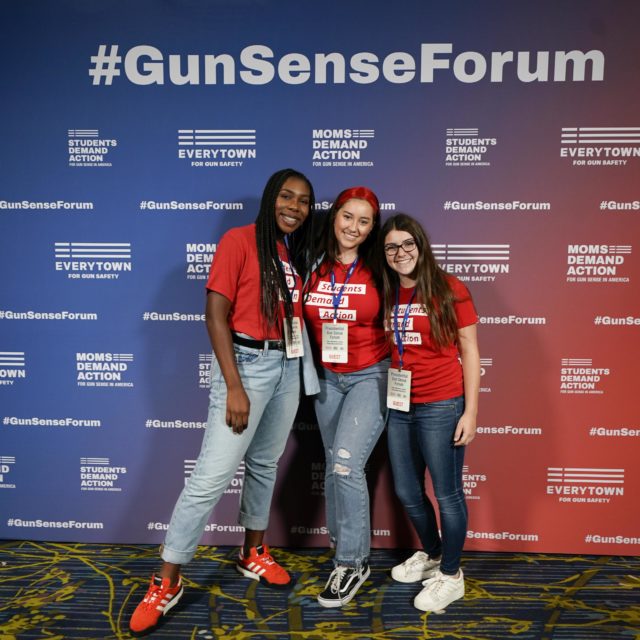 Alanna Miller and two other Students Demand Action volunteers pose for a photo in front of a step and repeat background at the Gun Sense Forum