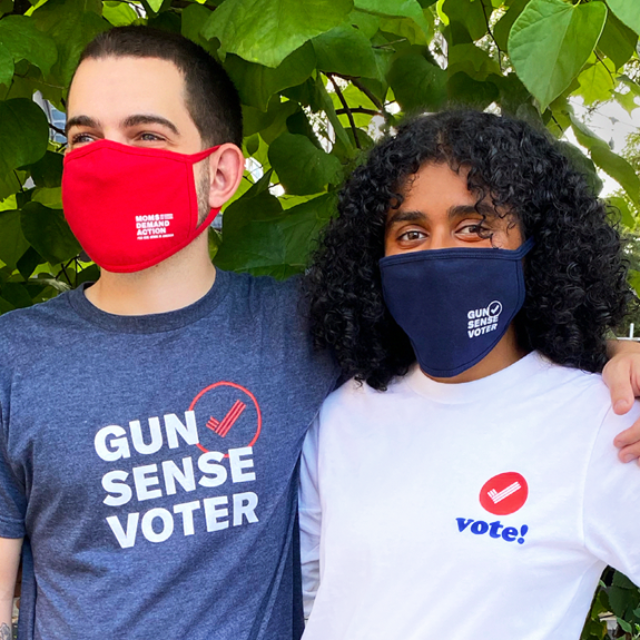 Two people stand together while wearing Gun Sense Voter merchandise including masks and tshirts