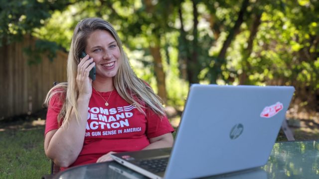 A woman in a Moms Demand Action shirt sits outside while making a call on her phone