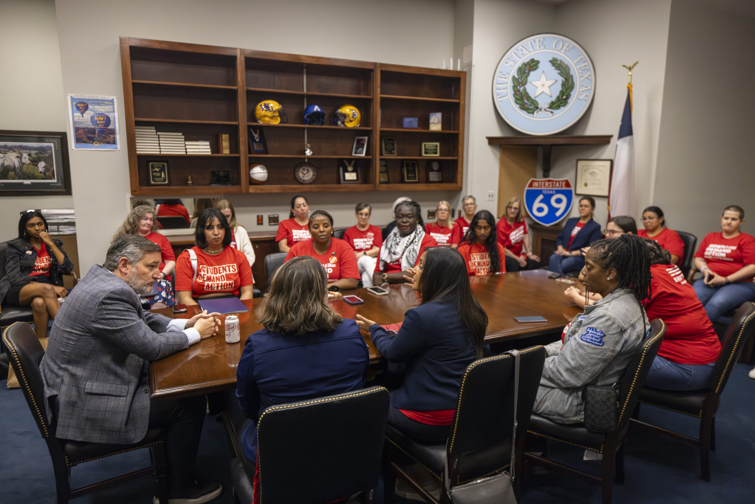 A total of 21 Moms Demand Action and Students Demand Action volunteers meet with Sen. Ted Cruz in his office in D.C. They all sit at a brown wood table and all except Cruz wear red branded t-shirts. Cruz wears a grey suit and a white collared shirt. 