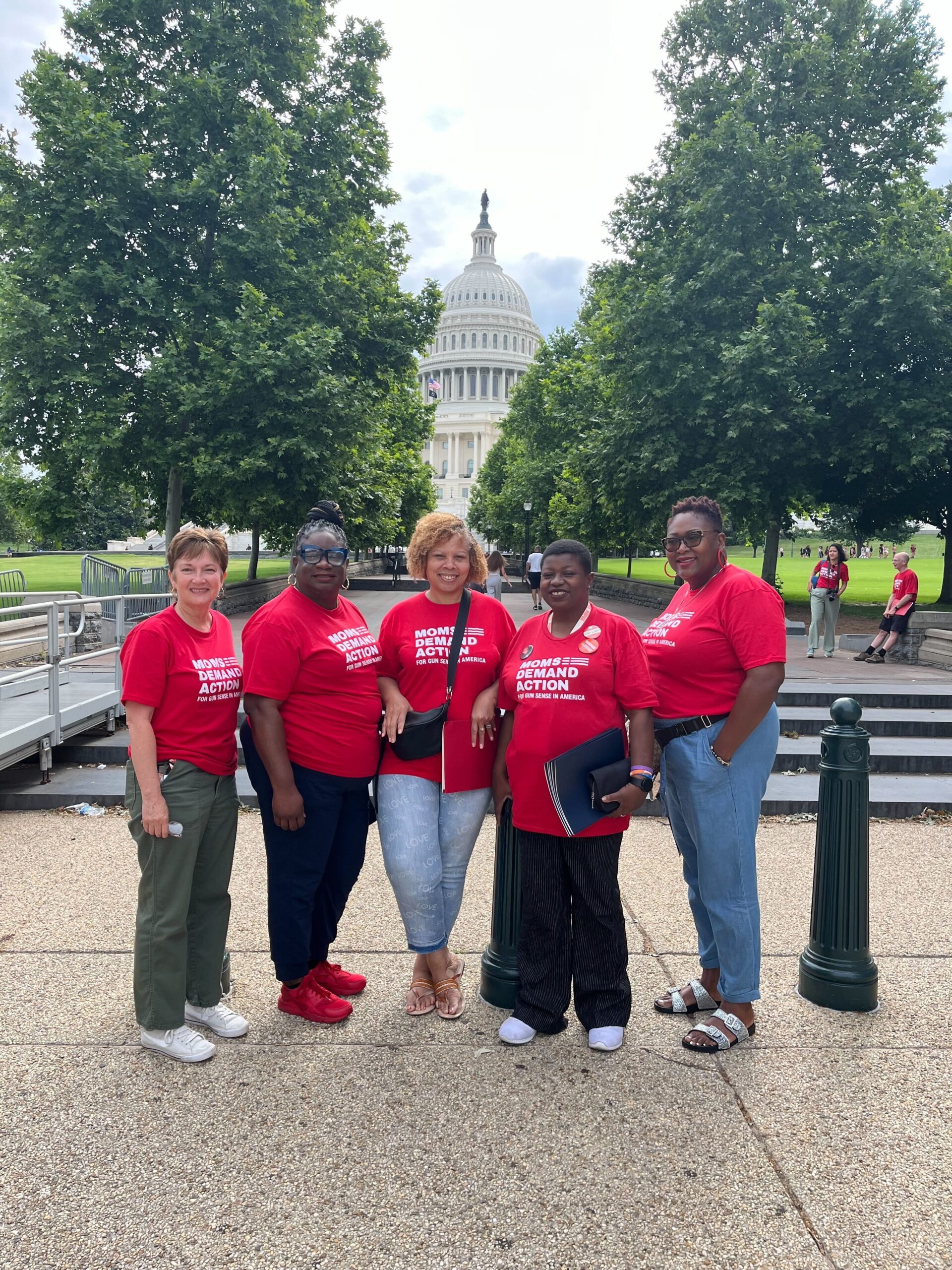Five volunteers from Alabama pose for a photo in front of the Capitol Building. Shade trees line the path that extends directly behind them; the Capitol building is visible in the center back of the photo.