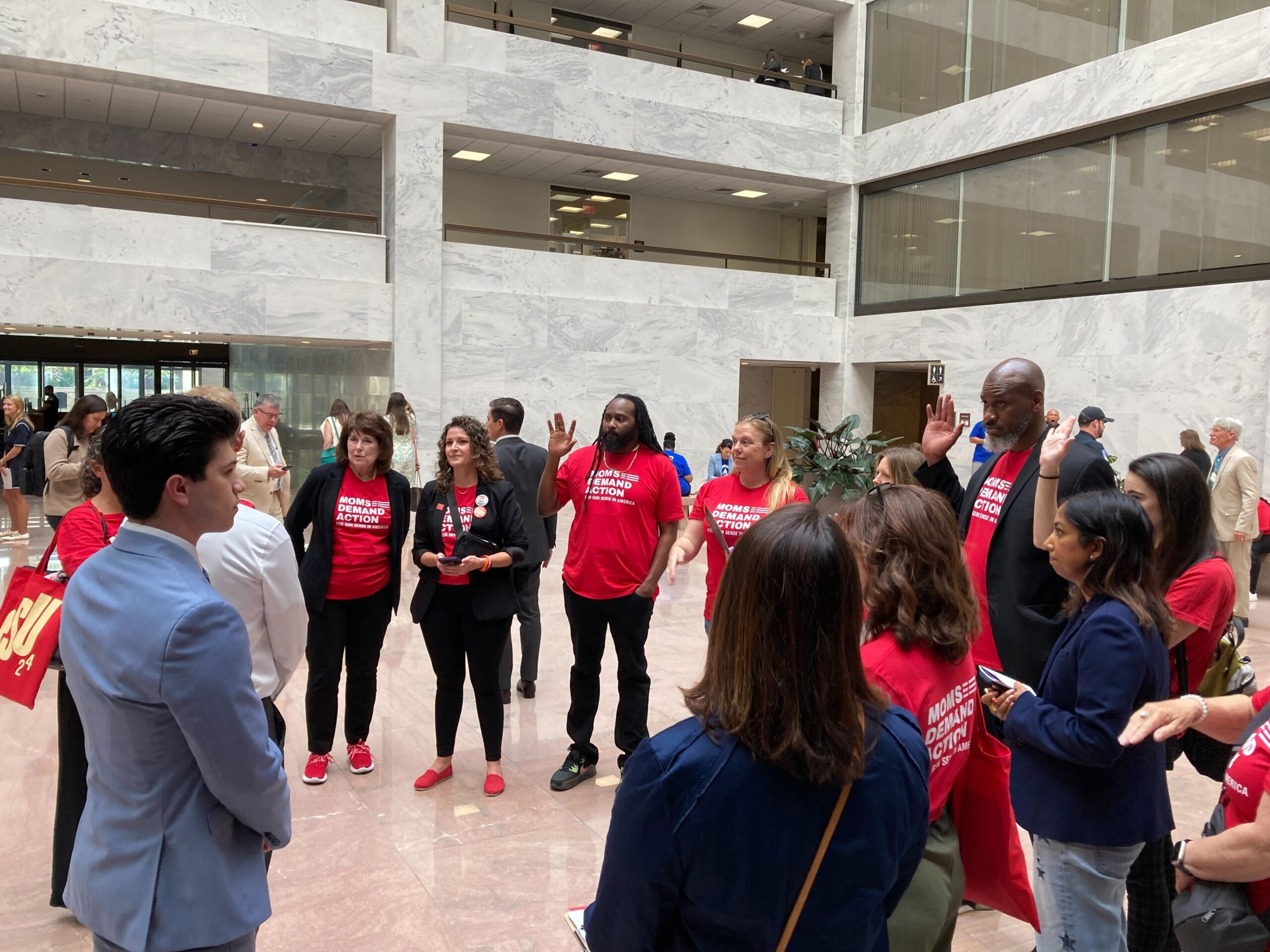At least 14 Moms Demand Action volunteers stand in a semi-circle around Rep. Mike Garcia's staffer. The staffer wears a light blue suit jacket and a white collared shirt; the California volunteers wear red branded tees. 