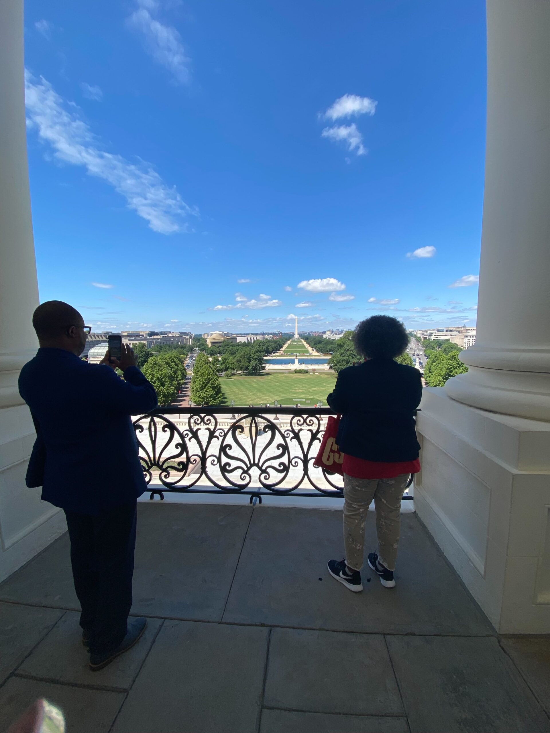 The two volunteers from Louisiana stand on the balcony behind the Speaker's Office. They stare out across the Capitol Steps and toward the National Mall and Washington Monument. On the right of the photo, the male volunteer is taking a picture of the view; on the left, the female volunteer is looking out at the Mall.