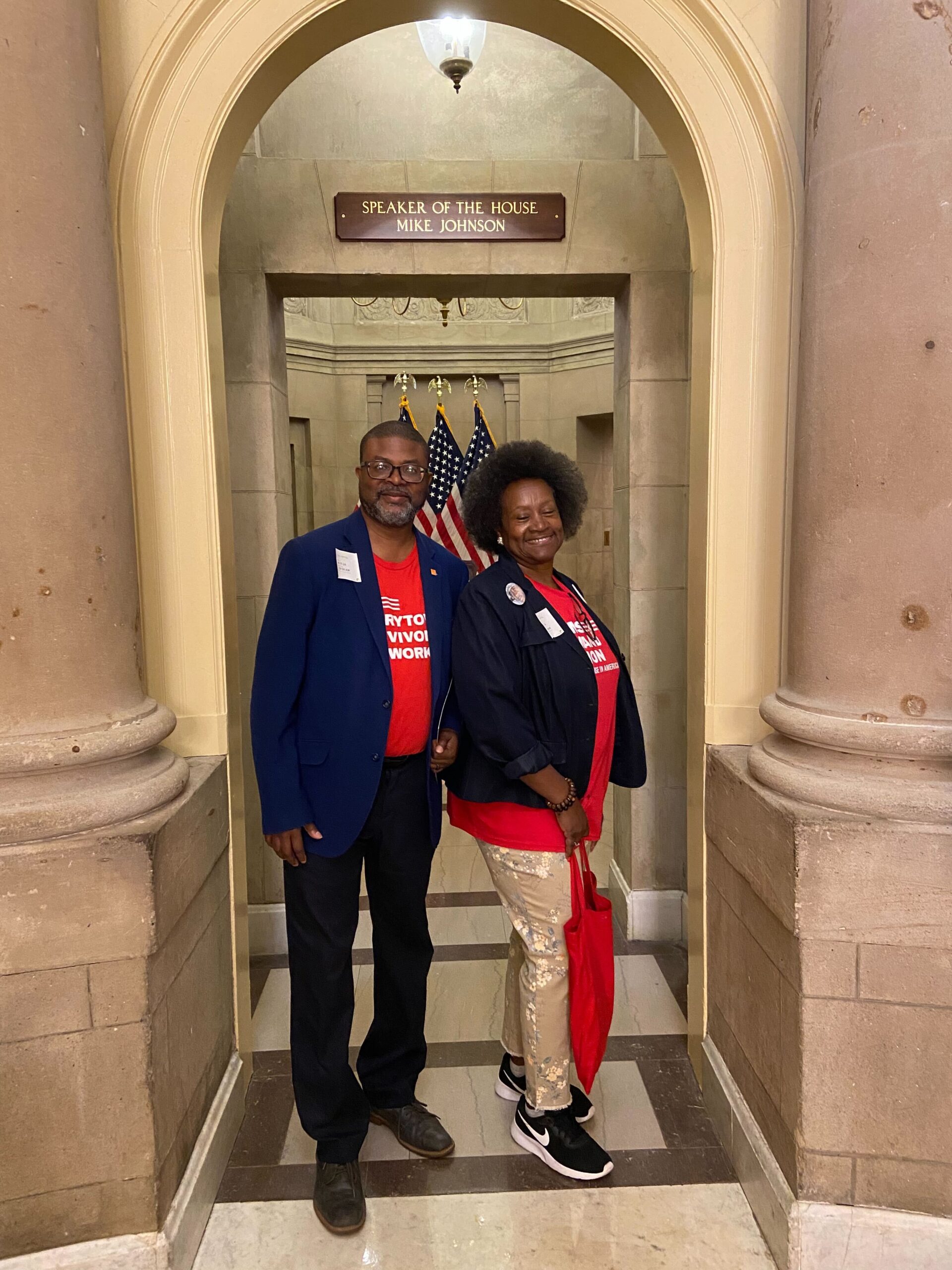 Two volunteers from Louisiana pose in front of Speaker of the House Mike Johnson's office in the D.C. Capitol. Both volunteers—a man on the right, and a woman on the left—are Black; they both wear red branded shirts and dark jackets overtop of the tees.