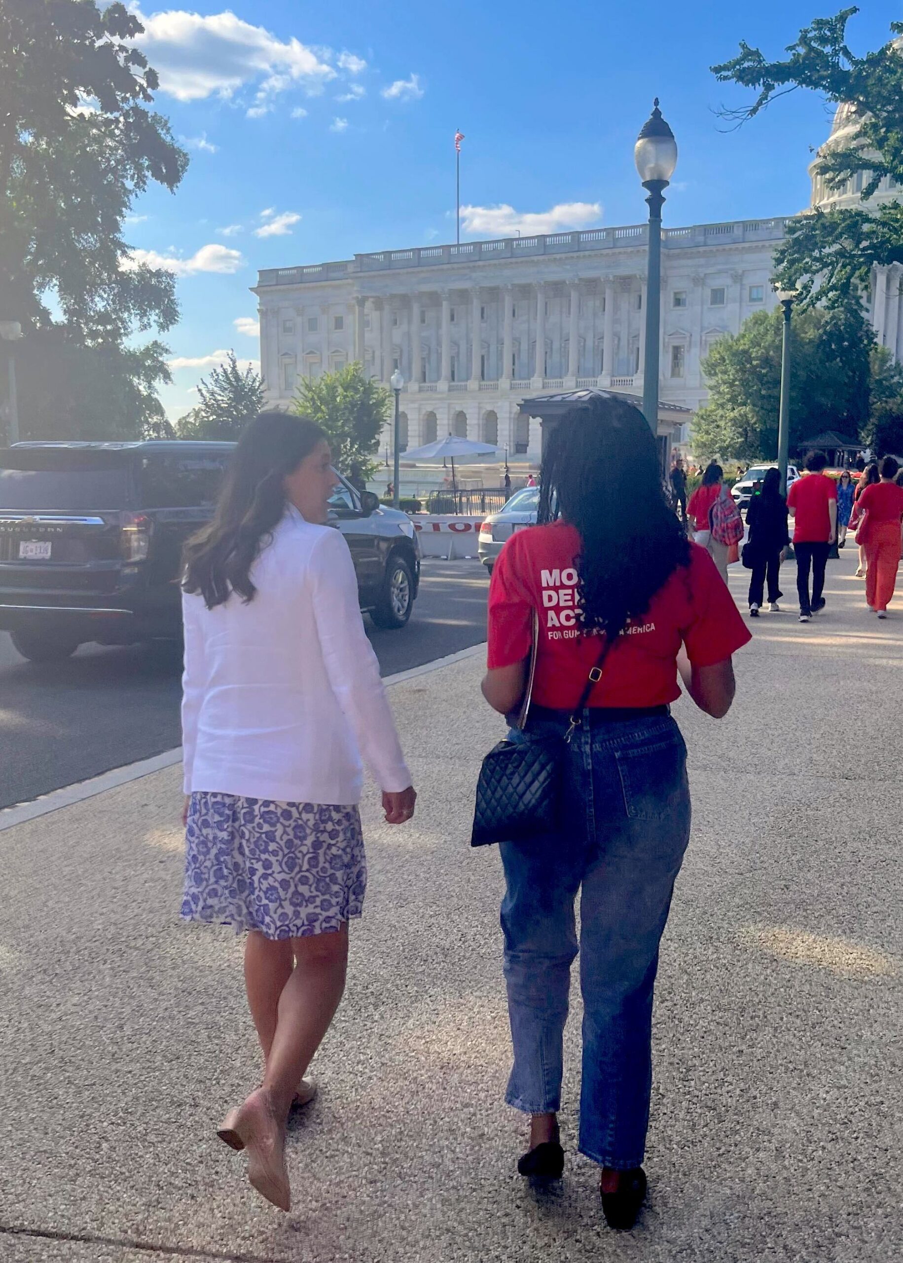 Two people are pictured from the back, walking toward the Capitol Building. Rep. Stephanie Bice has long dark hair that is curled; she wears a white linen blazer and a white-and-blue floral patterned dress. JeKia Harrison is a Black woman with dark braids, a red Moms Demand Action t-shirt, blue jeans, and a dark purse. 