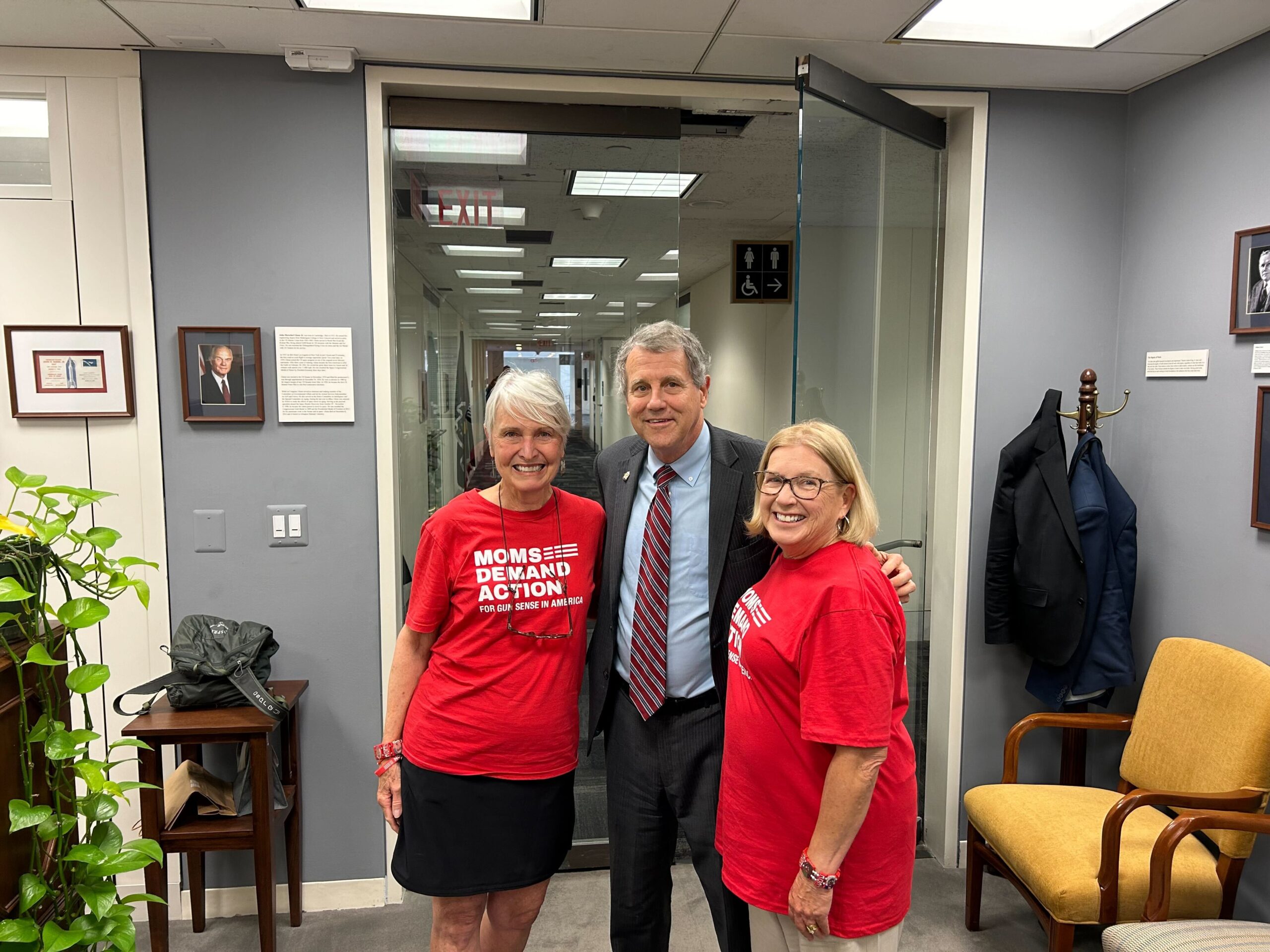 Two volunteers from Ohio pose for a photo with Rep. Sherrod Brown. Both the volunteers wear red Moms Demand Action tees; Rep Brown stands between them. He wears a dark suit jacket, a light blue collared shirt, and a red and blue striped tie.