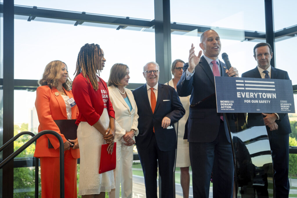 Hakeem Jeffries speaks at an Everytown-branded podium. From left to right, Lucy McBath, Angela Ferrell-Zabala, Nancy Pelosi, John Feinblatt, Hakeem Jeffries, and Chris Murphy stand in a row. The others are watching Jeffries speak as he holds a microphone in his left hand and gestures with his right.